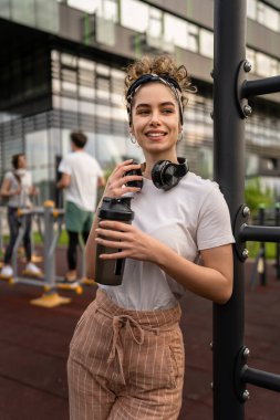 Beyaz kadın açık hava antrenmanı sırasında fren yapıyor açık hava spor salonunda barlarda dinleniyor el sıkışma aleti ile su içiyor ya da ek olarak gülümsüyor.
