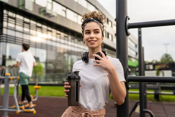 Beyaz kadın açık hava antrenmanı sırasında fren yapıyor açık hava spor salonunda barlarda dinleniyor el sıkışma aleti ile su içiyor ya da ek olarak gülümsüyor.
