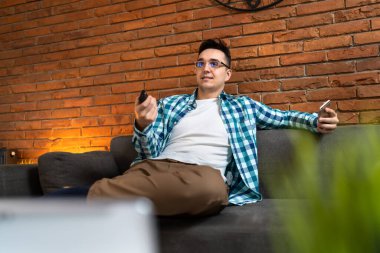 Yetişkin bir erkek, evdeki kanepede oturup uzaktan kumandayla uzaktan kumanda kullanan beyaz bir erkek.