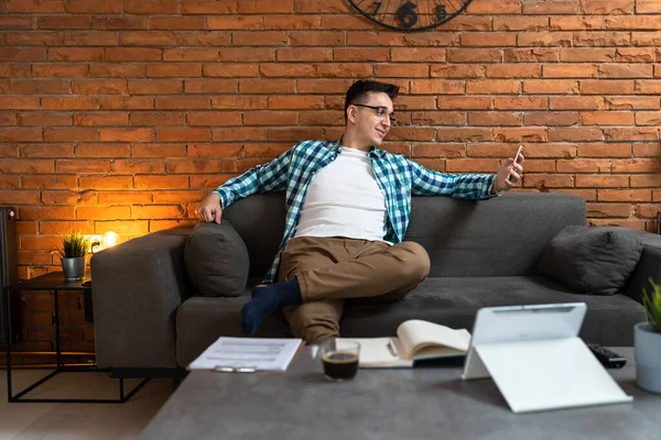 Genç, yetişkin bir erkek, evdeki kanepede oturup internetten sohbet etmek için cep telefonunu kullanarak fren yapıyor. Ya da mutlu bir gülümseme.