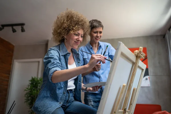 Two women female friends or woman with her mentor art professor caucasian mature sisters teach and learn how to paint helping during the painting ont the easel at home in bright room copy space