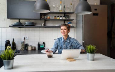 Yetişkin bir kadın mutfakta kahve içerken dijital tablet kullanıyor. Günlük posta ya da günlük işleri kontrol ediyor. Gerçek insanlar işe hazırlanıyor.