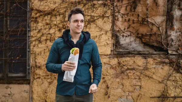 秋の春や冬の日に屋外に立つ1人の男古い壁の前で屋外を待っている間に足で食べるサンドイッチの観光客を保持実際の人々はスペースをコピーしますファーストフードの概念 — ストック写真