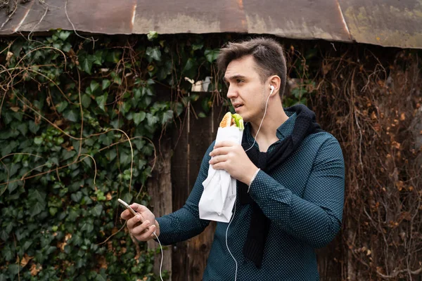 秋の春や冬の日に屋外に立つ1人の男古い壁の前で屋外を待つ間に食べるサンドイッチの観光客を保持する携帯電話を使用します — ストック写真