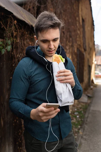 秋の春や冬の日に屋外に立つ1人の男古い壁の前で屋外を待つ間に食べるサンドイッチの観光客を保持する携帯電話を使用します — ストック写真