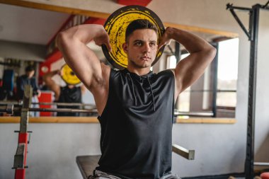 Genç, yetişkin, beyaz bir erkek ağırlık plakası tutarak spor salonunda oturuyor ve omuzlarının üzerinde antrenman yapıyor. Gerçek insanlar, amatör halter konsepti.