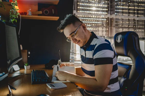 Genç bir yetişkin beyaz erkek, masa ve bilgisayar başında otururken kripto para kazanma işi ve ticaretine yaptığı yatırımı hesaplıyor.