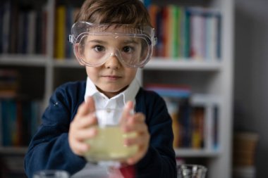 Beş yaşında, koruyucu gözlük takan beyaz bir bilim adamı kitaplığın önünde kameranın önünde durup öğrenme ve eğitim konseptine bakıyor.