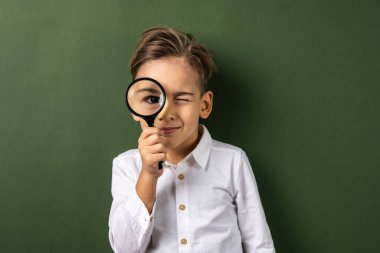 Yeşil arka plan duvarının önünde elinde mercek büyüteci olan bir çocuk kameraya bakıyor. Beyaz gömlekli, bilim ve eğitim konsepti giymiş.