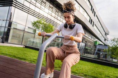 Gündüz bina önünde kahverengi saçlı, modern beyaz bir kadın spor salonunda bisiklet eğitmeni simülatöründe sağlıklı yaşam tarzı konseptini taklit ediyor.