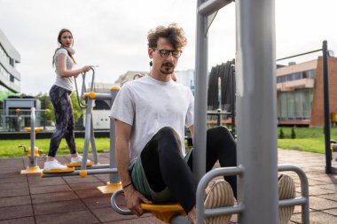 Açık spor salonunda erkek antrenmanı, arka planda genç bir kadınla bacak egzersizi, erkek arkadaş, erkek arkadaş spor ve eğlence gibi sağlıklı yaşam tarzı konsepti fotokopi alanı.