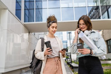 Modern üniversite binasının önünde cep telefonu kullanarak notlar ve kağıt belgelerle yürüyen iki kadın öğrenci ellerinde cep telefonu mesajları okurken mutlu bir gülümseme ile konuşuyorlar.