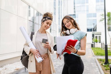 İki kadın öğrenci yaz günü üniversite binasının önünde birlikte yürürken notlar tutarak sınav ya da ders hakkında konuşuyorlar.