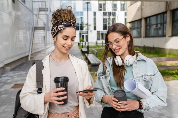 两名女学生拿着便条和纸质文件走在现代大学大楼前 一边拿着手机短信一边谈笑风生 — 图库照片