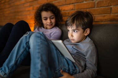 Küçük beyaz çocuk evde dijital tablet kullanarak kız kardeşinin yanında oturuyor.