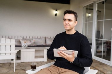 Genç bir beyaz adam, elinde dijital tabletle evinin önündeki sandalyede oturuyor ve şöyle düşünüyor: