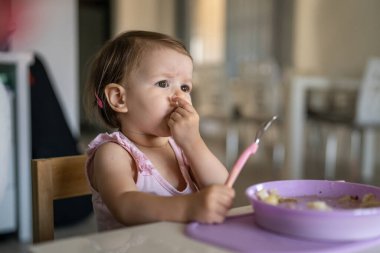 Bir çocuk, küçük beyaz bir bebek, evde masada tek başına yemek yiyor. Gerçek insanlar, erken dönem çocuk gelişimi konseptini taklit ediyor.