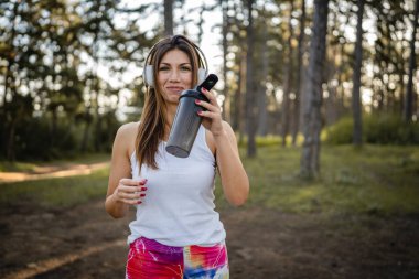 One woman happy caucasian female standing in the forest or park in woods with supplement shaker and headphones during training in nature taking a brake water drink and hydration real people copy space