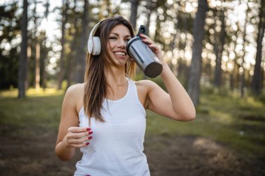 One woman happy caucasian female standing in the forest or park in woods with supplement shaker and headphones during training in nature taking a brake water drink and hydration real people copy space