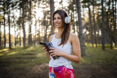 One woman happy caucasian female standing in the forest or park in woods with supplement shaker and headphones during training in nature taking a brake water drink and hydration real people copy space