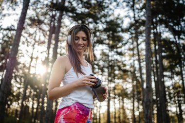 One woman happy caucasian female standing in the forest or park in woods with supplement shaker and headphones during training in nature taking a brake water drink and hydration real people copy space