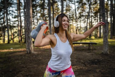 One woman adult caucasian female athlete using kettlebell girya weight during training in the forest park woods in summer day with headphones happy smile brunette health and fitness concept copy space