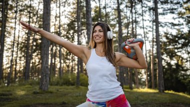 One woman adult caucasian female athlete using kettlebell girya weight during training in the forest park woods in summer day with headphones happy smile brunette health and fitness concept copy space