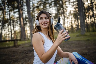 One woman happy caucasian female sitting in the forest or park in woods with supplement shaker and headphones during training in nature taking a brake water drink and hydration real people copy space