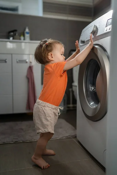 One girl small caucasian toddler child daughter standing at the washing machine in the toilet opening or closing the door examine and learn early development and growing up mischief concept copy space