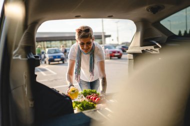 Olgun bir beyaz kadın güneşli bir günde arabasının arkasına sebze dolu bir kutu koyuyor. Süpermarketin önündeki otoparkta ya da markette. Gerçek insanların fotokopi çektikleri yerlerde.