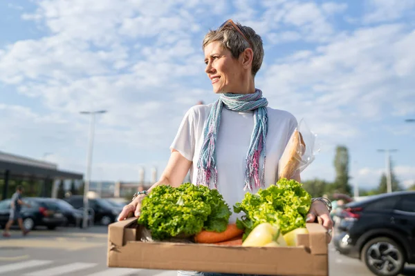 One woman mature caucasian female standing in the parking lot in front of supermarket grocery store holding box with food and vegetables in sunny day happy smile healthy eating vegan concept