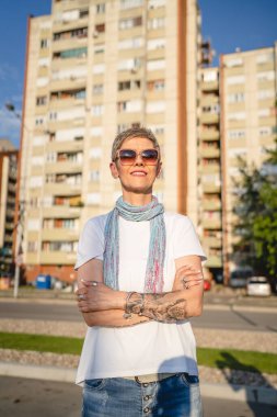 Güneşli yaz gününde, açık havada duran, gözlüklü, kısa gri saçlı, mutlu, kendine güvenen, kameraya bakan beyaz tişörtlü, olgun bir kadın.