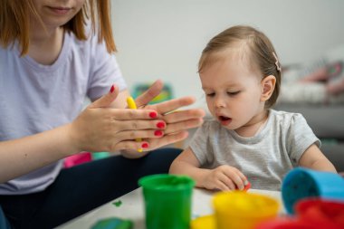 One girl small caucasian toddler child playing with colorful plasticine on the table at home with her mother woman childhood and growing up development concept copy space side view