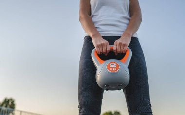 Beyaz tenli kadın sporcunun orta kesiminde kettlebell girya plastik ağırlığı tutarken yaz akşamı antrenman sırasında stadyumda ayakta duruyor.