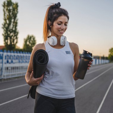 Yetişkin bir beyaz kadın sporcu stadyumda koşuyor. Takviye ile koşuyor ve kulaklıklı yoga minderi takıyor. Yazın açık havada eğitime hazır.