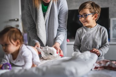 Beyaz çocuk, küçük çocuk ve annesi, erkek ya da kız kardeşlerine bakıyorlar, üzerlerini değiştirip evde yatıyorlar. Gerçek insanlar, aile ve bağ kurma kavramı.