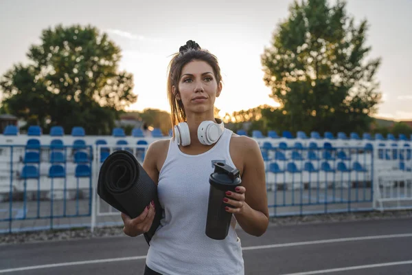 one woman adult caucasian female athlete standing on stadium on running track with supplement shaker and yoga mat with headphones ready for training outdoor in summer evening real people copy space