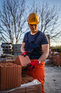 Bir inşaat işçisi, güneşli yaz gününde kil portakalı blokları depo ya da şantiye alanlarını alıp taşıyor ya da tutuyor.