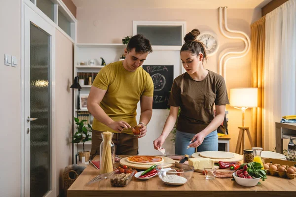 Two people young adult couple man and woman husband and wife or boyfriend and girlfriend preparing pizza food at home helping each other cutting and add ingredients and spices real people copy space