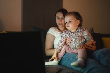 Küçük beyaz çocuk, küçük kız bebek anne ve kız geceleri karanlık bir odada kanepe yatağında oturup çizgi film ya da dizüstü bilgisayar filmleri izliyorlar.