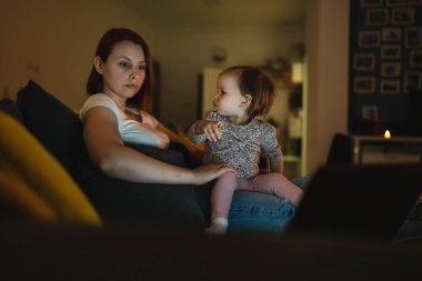 Küçük beyaz çocuk, küçük kız bebek anne ve kız geceleri karanlık bir odada kanepe yatağında oturup çizgi film ya da dizüstü bilgisayar filmleri izliyorlar.