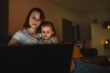 Küçük beyaz çocuk, küçük kız bebek anne ve kız geceleri karanlık bir odada kanepe yatağında oturup çizgi film ya da dizüstü bilgisayar filmleri izliyorlar.