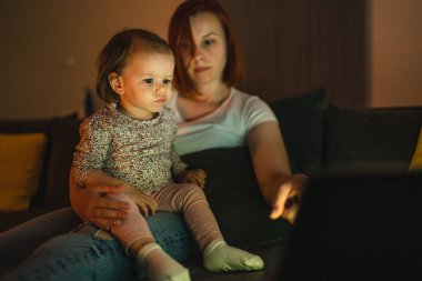 Small caucasian child little girl toddler mother and daughter sitting in dark room at night at home on sofa bed watching cartoons or movies on laptop computer real people family growing up copy space