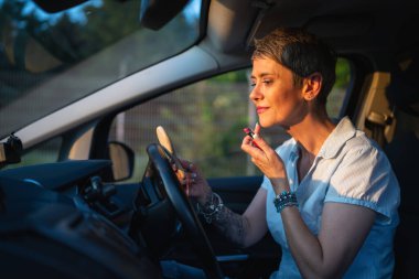 Yetişkin bir beyaz kadın iş kadını arabada oturuyor yüzüne ruj sürüyor makyaj yapıyor ve yaz akşamı bekliyor gerçek insanlar uzay grisini taklit ediyor.