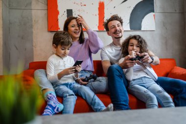 Bir grup Kafkasyalı aile üyesi, anne-baba ve çocuklar evde kanepede oturup oyun konsolu oynarken oyun konsolu ya da kumanda çubuğu veya gerçek insanlar konseptini kullanıyorlar.