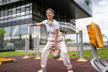 Gündüz bina önünde kısa saçlı, modern, olgun bir beyaz kadın spor salonunda ağırlık sallıyor sağlıklı yaşam tarzı konseptini taklit ediyor.