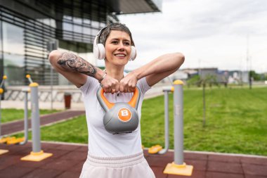 Bir kadın, modern olgun, beyaz kadın. Kısa saçlı, bina önünde günün ilerleyen saatlerinde spor salonunda kettlebell ağırlığı ile spor salonu sağlıklı yaşam tarzı konseptini taklit ediyor.