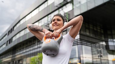 Bir kadın, modern olgun, beyaz kadın. Kısa saçlı, bina önünde günün ilerleyen saatlerinde spor salonunda kettlebell ağırlığı ile spor salonu sağlıklı yaşam tarzı konseptini taklit ediyor.