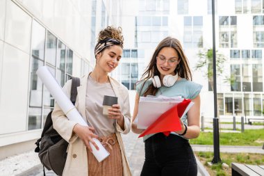 İki kadın öğrenci yaz günü üniversite binasının önünde birlikte yürürken notlar tutarak sınav ya da ders hakkında konuşuyorlar.