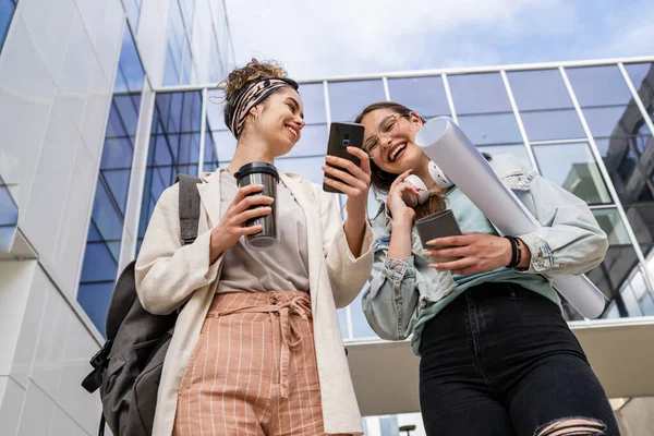two women female students walk in front of modern university building with notes and paper documents using mobile phone discuss talk happy smile while holding mobile phone reading sms text messages
